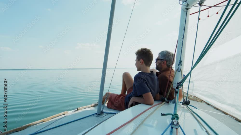 Modern mature man and his teen son sitting relaxed on board the sailboat talking about their travel Modern mature man and his teen son sitting relaxed on board the sailboat talking about their travel