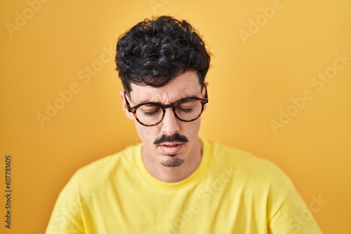 Hispanic man wearing glasses standing over yellow background with hand on stomach because indigestion, painful illness feeling unwell. ache concept.