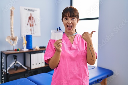 Young brunette woman working at rehabilitation clinic showing id pointing thumb up to the side smiling happy with open mouth