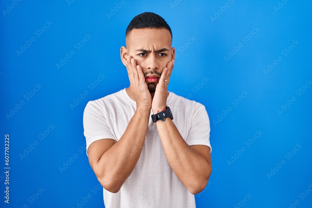 Young hispanic man standing over blue background tired hands covering face, depression and sadness, upset and irritated for problem