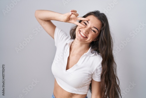 Young teenager girl standing over white background doing peace symbol with fingers over face, smiling cheerful showing victory