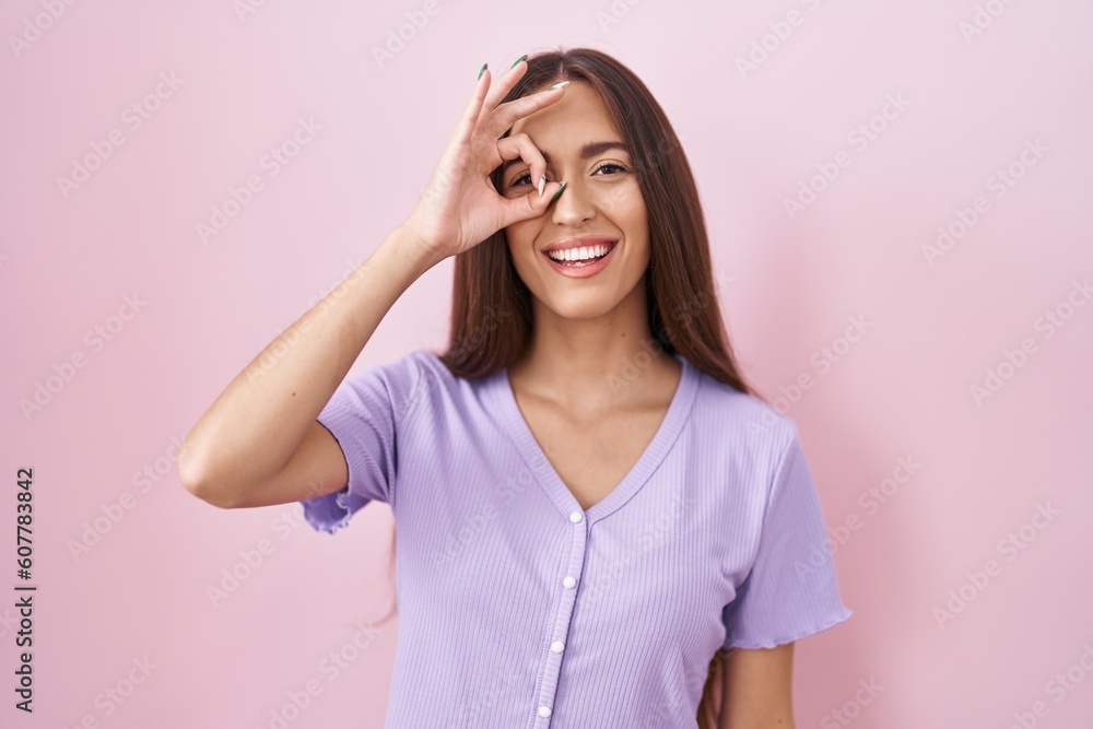 Young hispanic woman with long hair standing over pink background doing ok gesture with hand smiling, eye looking through fingers with happy face. Young hispanic woman with long hair standing over pink background doing ok gesture with hand smiling, eye looking through fingers with happy face.