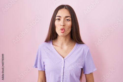 Young hispanic woman with long hair standing over pink background making fish face with lips, crazy and comical gesture. funny expression.