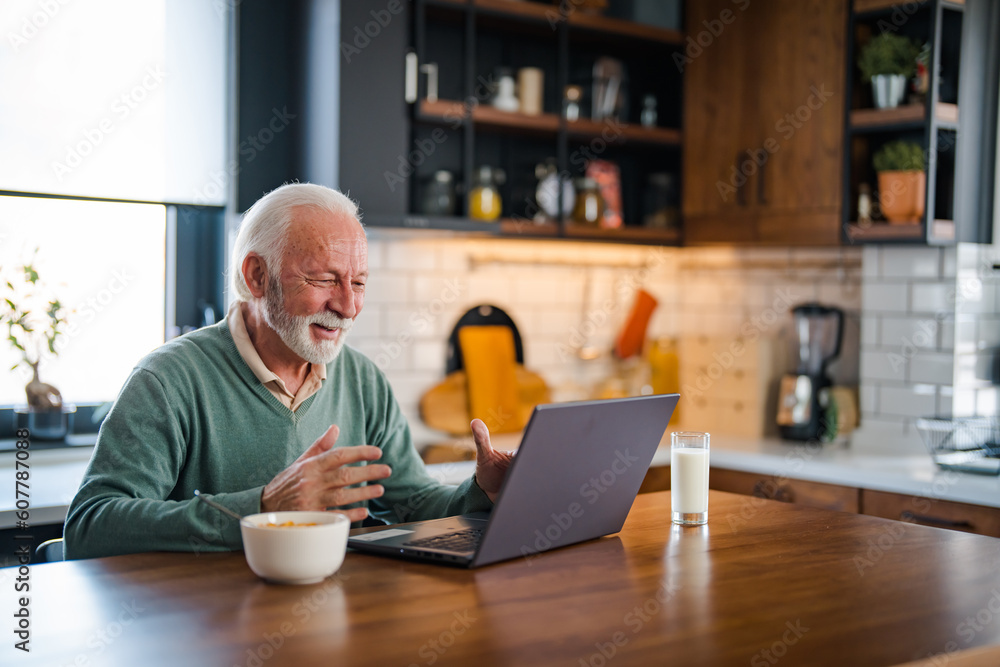 One senior businessman standing in the kitchen and working on the laptop while drinking coffee. Grey-haired bearded senior man eyeglasses looking concentrated One senior businessman standing in the kitchen and working on the laptop while drinking coffee. Grey-haired bearded senior man eyeglasses looking concentrated