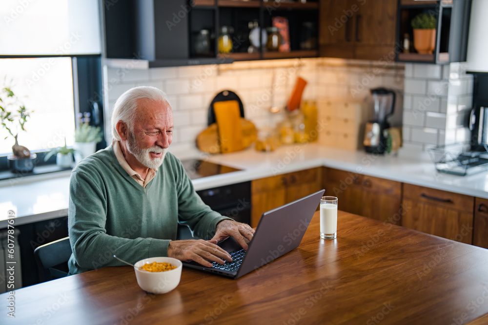 Man with computer at home in modern kitchen. Senior man connected on internet with laptop at home. Remote Work On Retirement. Smiling Senior Man Using Laptop In Kitchen At Home Man with computer at home in modern kitchen. Senior man connected on internet with laptop at home. Remote Work On Retirement. Smiling Senior Man Using Laptop In Kitchen At Home