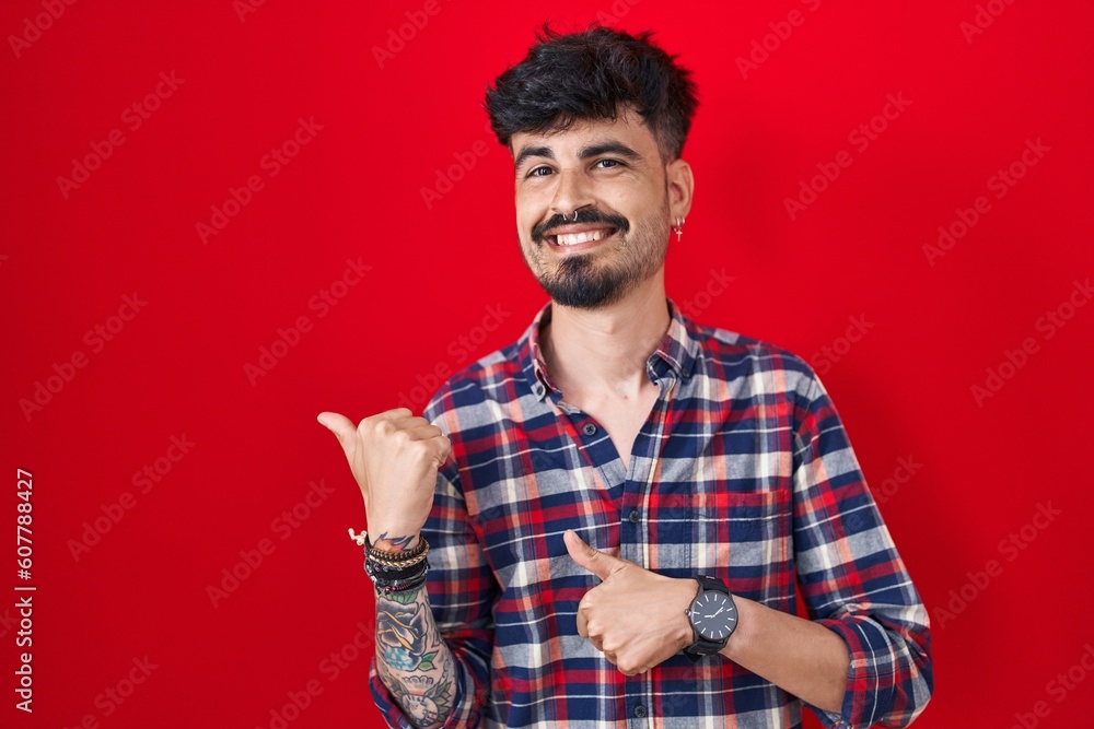 Young hispanic man with beard standing over red background pointing to the back behind with hand and thumbs up, smiling confident Young hispanic man with beard standing over red background pointing to the back behind with hand and thumbs up, smiling confident