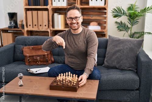 Middle age caucasian man playing chess sitting on the sofa smiling happy pointing with hand and finger