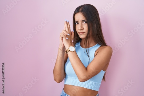 Young brunette woman standing over pink background holding symbolic gun with hand gesture, playing killing shooting weapons, angry face