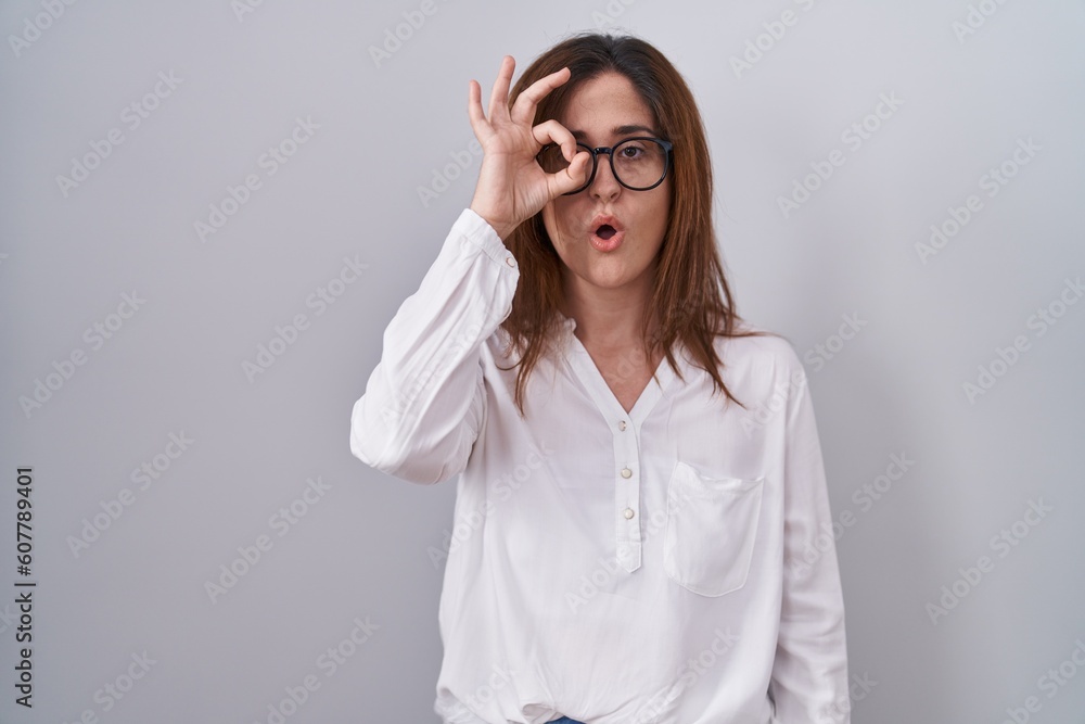 Brunette woman standing over white isolated background doing ok gesture shocked with surprised face, eye looking through fingers. unbelieving expression. Brunette woman standing over white isolated background doing ok gesture shocked with surprised face, eye looking through fingers. unbelieving expression.