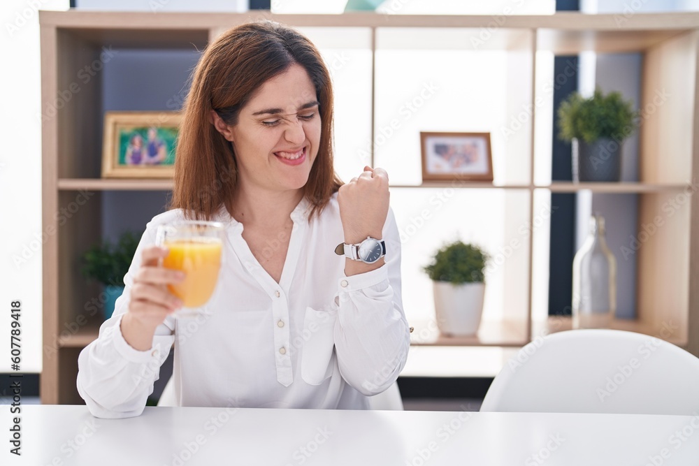 Brunette woman drinking glass of orange juice very happy and excited doing winner gesture with arms raised, smiling and screaming for success. celebration concept. Brunette woman drinking glass of orange juice very happy and excited doing winner gesture with arms raised, smiling and screaming for success. celebration concept.