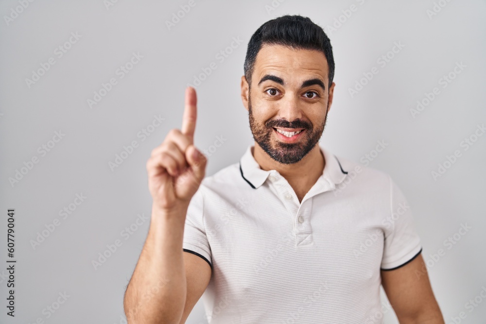 Young hispanic man with beard wearing casual clothes over white background showing and pointing up with finger number one while smiling confident and happy. Young hispanic man with beard wearing casual clothes over white background showing and pointing up with finger number one while smiling confident and happy.