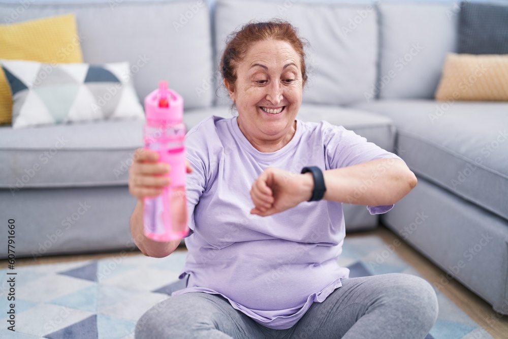 Fototapeta premium Senior woman sitting on floor drinking water looking watch at home