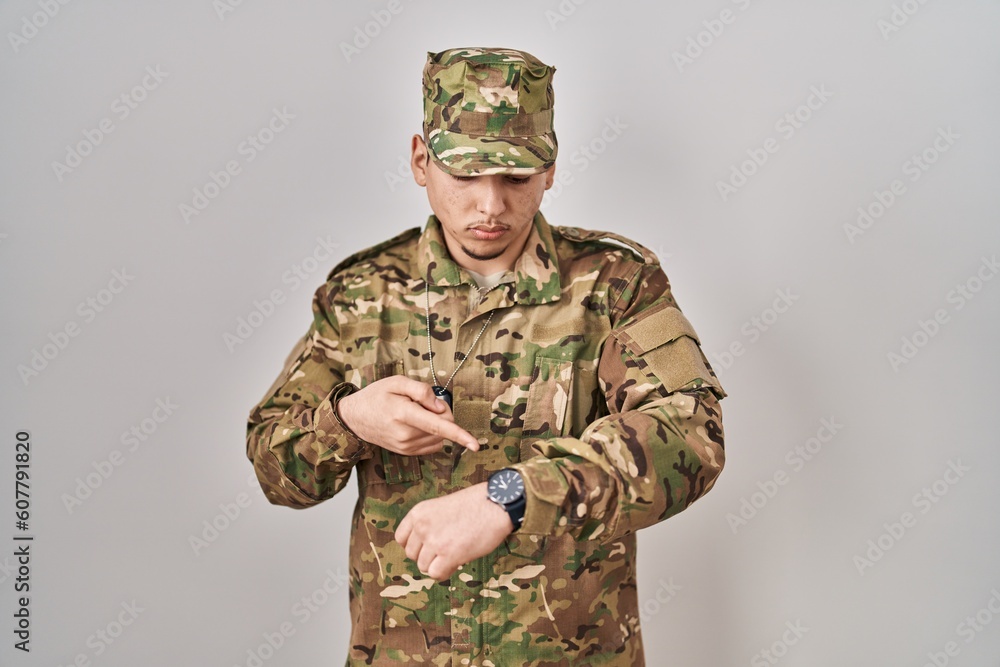 Young arab man wearing camouflage army uniform checking the time on wrist watch, relaxed and confident