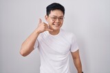Young asian man standing over white background smiling doing phone gesture with hand and fingers like talking on the telephone. communicating concepts.