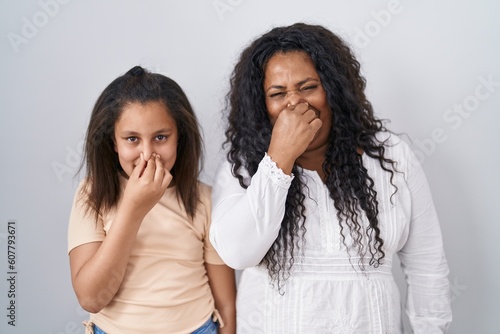 Mother and young daughter standing over white background smelling something stinky and disgusting, intolerable smell, holding breath with fingers on nose. bad smell