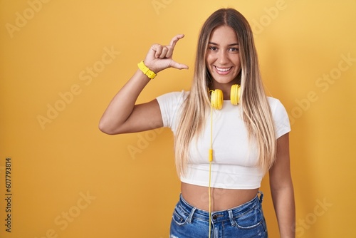 Young blonde woman standing over yellow background wearing headphones smiling and confident gesturing with hand doing small size sign with fingers looking and the camera. measure concept.