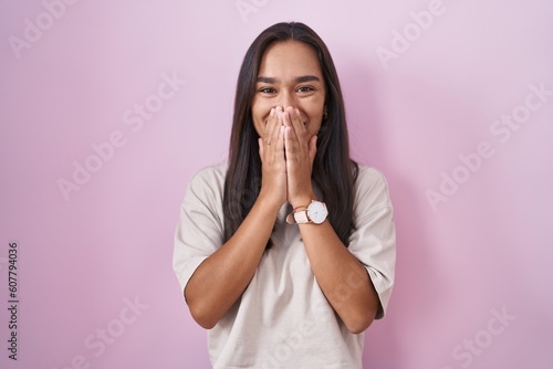 Young hispanic woman standing over pink background laughing and embarrassed giggle covering mouth with hands, gossip and scandal concept