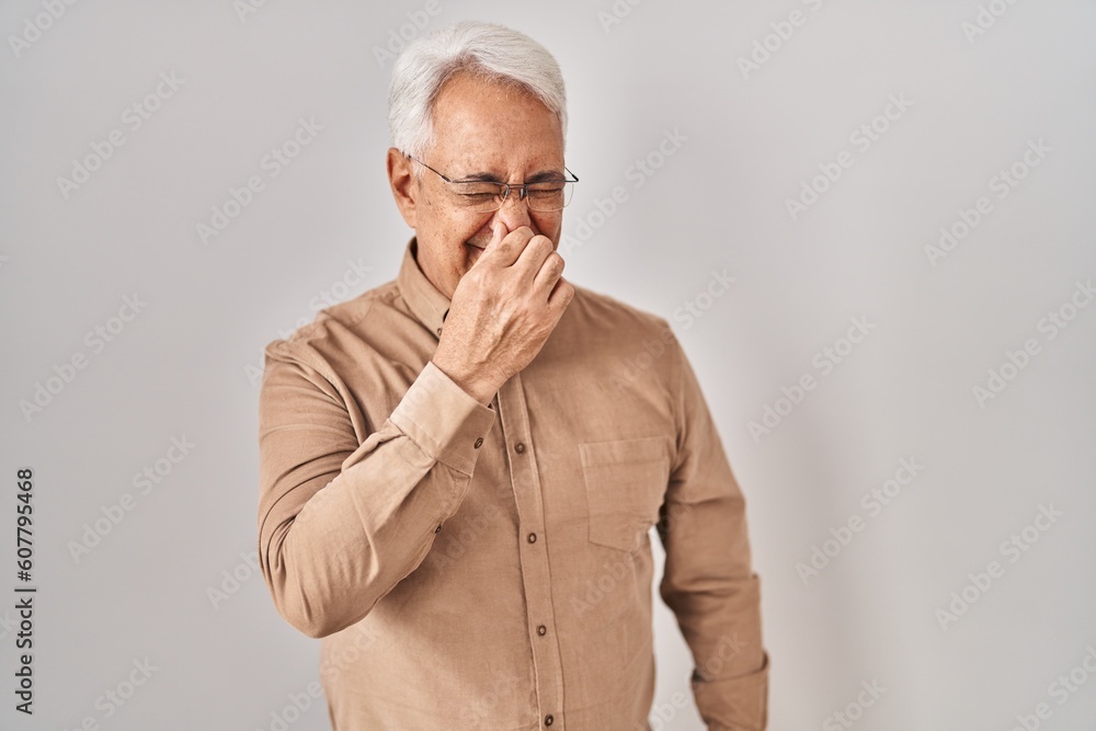 Hispanic senior man wearing glasses smelling something stinky and disgusting, intolerable smell, holding breath with fingers on nose. bad smell Hispanic senior man wearing glasses smelling something stinky and disgusting, intolerable smell, holding breath with fingers on nose. bad smell