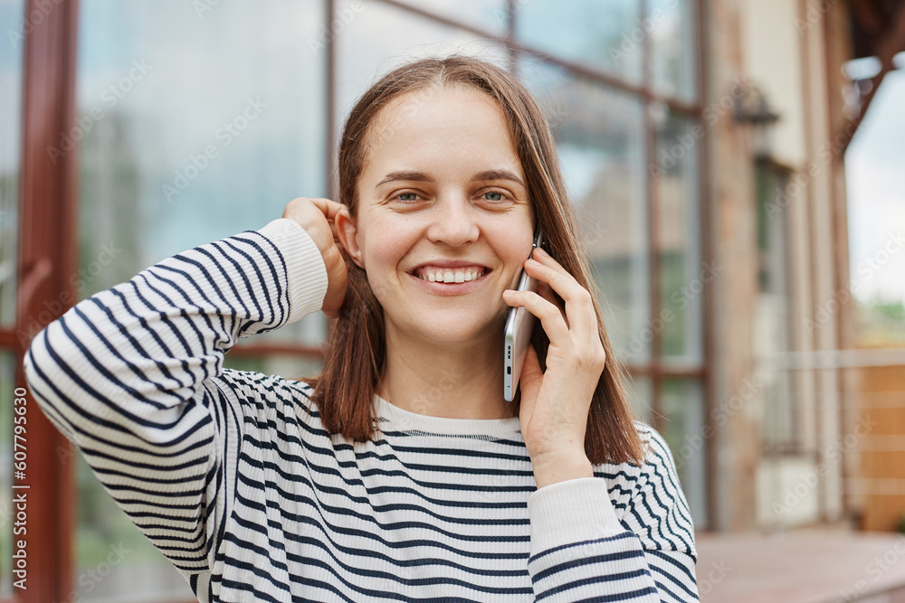 Cute brown haired looking at camera while talking on smartphone wearing striped shirt holding cell phone pleasant conversation while resting outdoor. Cute brown haired looking at camera while talking on smartphone wearing striped shirt holding cell phone pleasant conversation while resting outdoor.