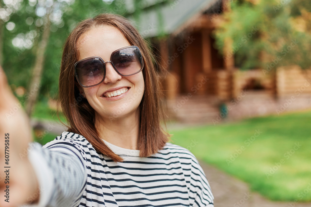 Smiling young dark haired girl making selfie photo in park with wooden house on background expressing positive emotions, POV, woman broadcasting livestream, having video call. Smiling young dark haired girl making selfie photo in park with wooden house on background expressing positive emotions, POV, woman broadcasting livestream, having video call.