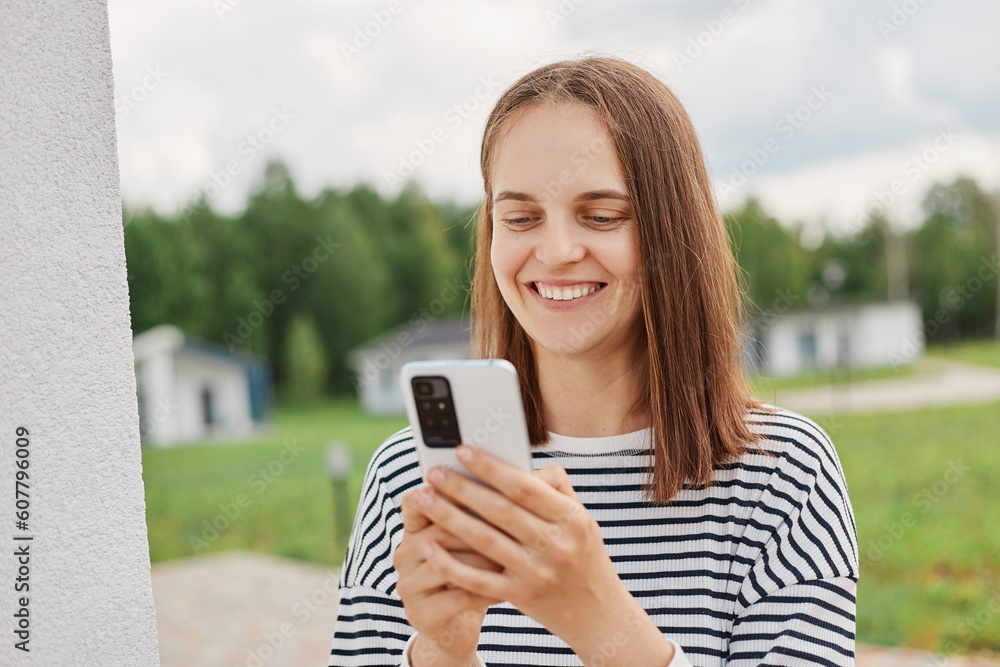 Portrait of cheerful joyful positive attractive female with brown hair standing in street wearing striped shirt holding cell phone in hands looking at device screen with smile. Portrait of cheerful joyful positive attractive female with brown hair standing in street wearing striped shirt holding cell phone in hands looking at device screen with smile.