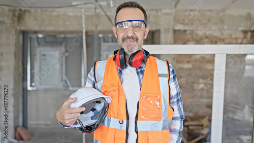 Middle age man builder smiling confident holding hardhat at construction site