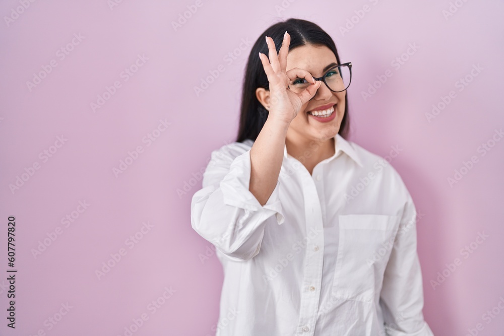 Young brunette woman standing over pink background doing ok gesture with hand smiling, eye looking through fingers with happy face. Young brunette woman standing over pink background doing ok gesture with hand smiling, eye looking through fingers with happy face.