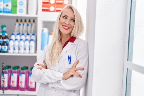 Young blonde woman pharmacist smiling confident standing with arms crossed gesture at pharmacy