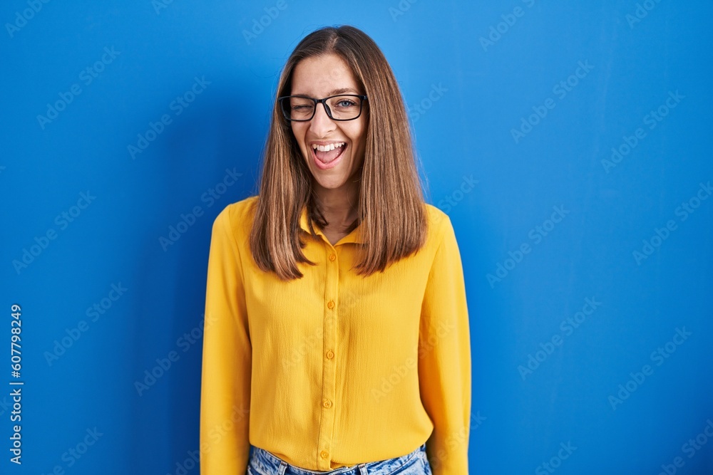 Young woman wearing glasses standing over blue background winking looking at the camera with sexy expression, cheerful and happy face. Young woman wearing glasses standing over blue background winking looking at the camera with sexy expression, cheerful and happy face.