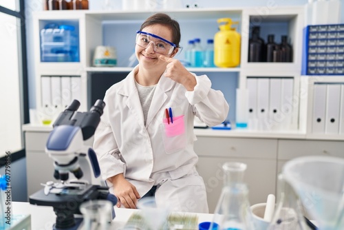 Hispanic girl with down syndrome working at scientist laboratory pointing with hand finger to face and nose, smiling cheerful. beauty concept