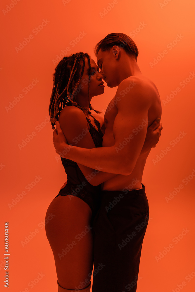 Naklejka premium side view of interracial passionate couple, shirtless man in black pants with muscular torso and sexy african american woman in lace bodysuit embracing on orange background with red lighting effect