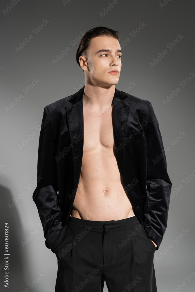 Fototapeta premium young, sexy and fashionable man wearing black silk blazer on shirtless muscular torso and looking away while posing with hands in pockets of pants on grey background
