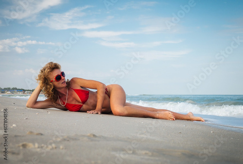 An adult redhead slender woman in a red swimsuit and red sunglasses lies on a sandy seashore and looks at the sea