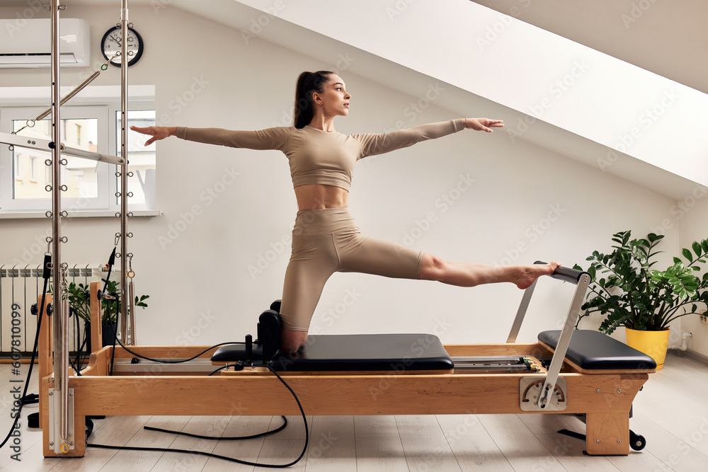 A young girl is doing Pilates on a reformer bed. A beautiful slender brunette woman in a beige bodysuit is doing body strengthening and stretching exercises.