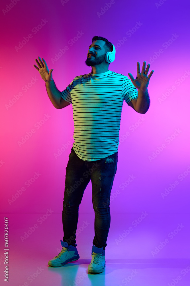 Full-length portrait of smiling bearded man in striped shirt listening to music in headphones against gradient pink blue studio background in neon light. Concept of human emotions, facial expression