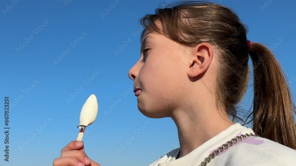 a happy brunette girl eats ice cream against the blue sky on a sunny summer day, a child appetitiously eats ice cream a happy brunette girl eats ice cream against the blue sky on a sunny summer day, a child appetitiously eats ice cream