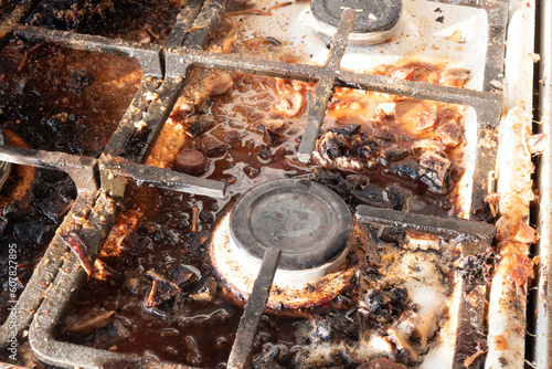 Dirty gas stove surface. Medium gas burner and cast iron grate of a gas oven surrounded by old leftovers of food and drinks. Top area surface and burner heads needs cleaning from burnt-on grease photo