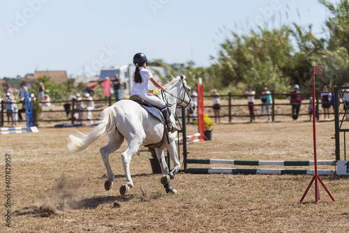 Equestrian sport - a little girl in uniform riding white horse at the ranch - jumping over the obsticles