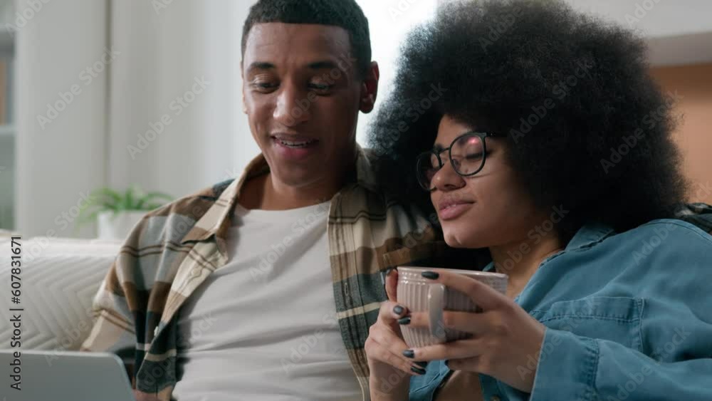 Happy family couple African American husband wife with cup of tea relaxing on sofa using laptop computer online shopping choose goods in internet shop store app discuss offers order delivery together Happy family couple African American husband wife with cup of tea relaxing on sofa using laptop computer online shopping choose goods in internet shop store app discuss offers order delivery together