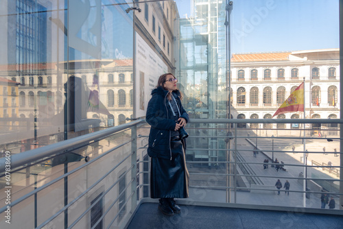 Young Latin woman tourist in warn clothes near window and looking out with professional photo camera in Reina Sofia museum in Madrid, Spain during daytime