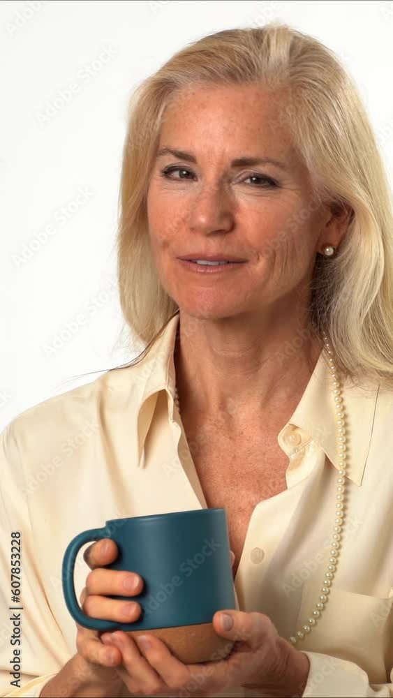 Vertical video closeup portrait of beautiful smiling happy blond mature woman holding cup drinking coffee or tea looking into camera on solid white background with copy space Vertical video closeup portrait of beautiful smiling happy blond mature woman holding cup drinking coffee or tea looking into camera on solid white background with copy space