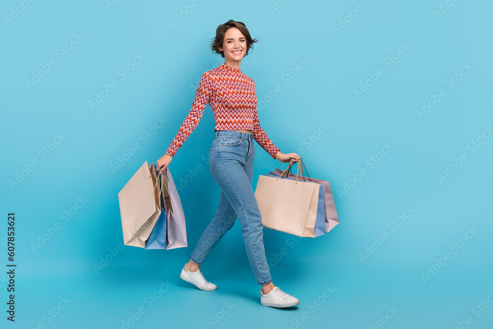Full length photo of excited shiny lady wear striped top walking holding shoppers isolated blue color background - obrazy, fototapety, plakaty 