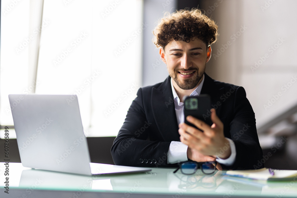 Smiling businessman chatting on phone in office. He is sitting at desktop with laptop