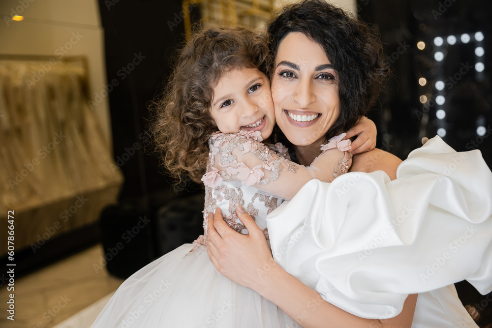 happy little girl in floral attire hugging tight her charming mother in white wedding dress with puff sleeves and ruffles while smiling and looking at camera together in bridal boutique