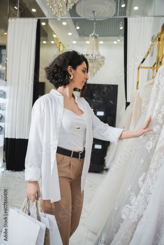 side view of stylish and happy middle eastern woman with brunette and wavy hair standing in beige pants with white shirt and holding shopping bags while choosing wedding dress in bridal salon