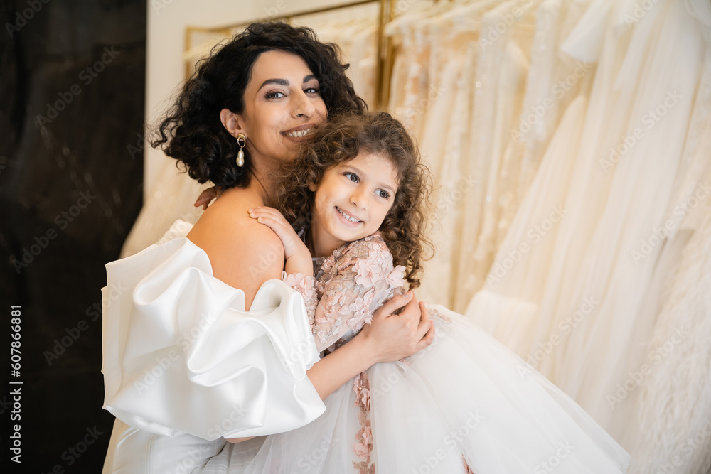 charming middle eastern bride with brunette hair in white wedding dress with puff sleeves and ruffles embracing positive daughter in bridal store, white tulle fabrics on blurred background