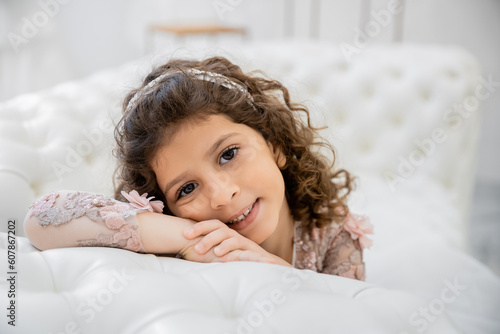 Wallpaper Mural portrait of positive middle eastern girl with brunette curly hair posing in floral dress and leaning on white couch inside of luxurious wedding salon, smiling kid, blurred background Torontodigital.ca