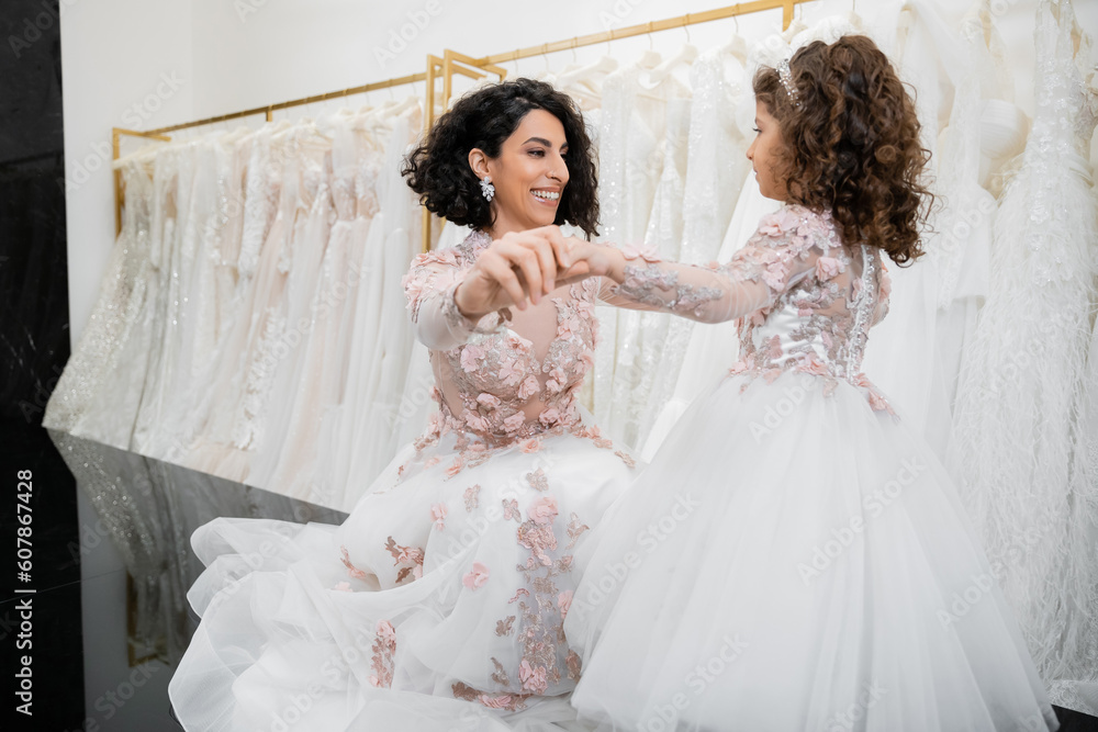 special moment, happy middle eastern bride in floral wedding gown sitting and holding hands with her little daughter in bridal salon around white tulle fabrics, bridal shopping, togetherness
