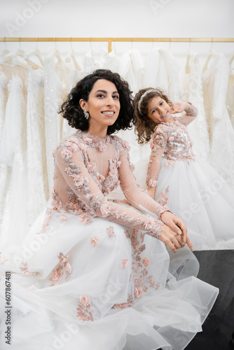 special moment, charming and happy middle eastern bride in floral wedding gown sitting next to her little daughter in bridal salon around white tulle fabrics, bridal shopping, togetherness
