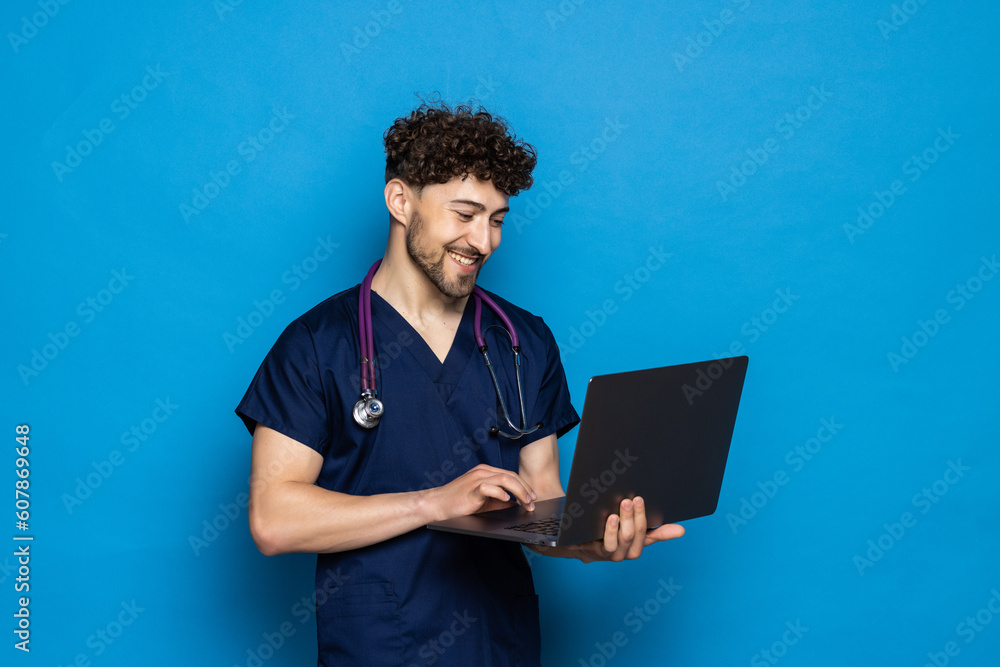 Doctor standing in the studio while working with laptop, isolated on blue background Doctor standing in the studio while working with laptop, isolated on blue background